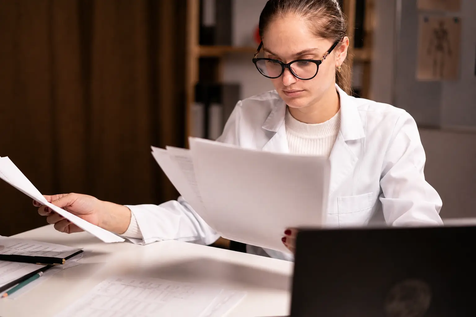 Clinician in a white coat reviewing paperwork at a desk with a laptop